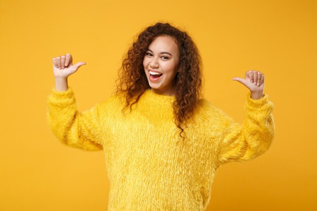 Photo cheerful funny young african american girl in fur sweater posing isolated on yellow orange wall background, studio portrait. people lifestyle concept. mock up copy space. pointing thumbs on herself.