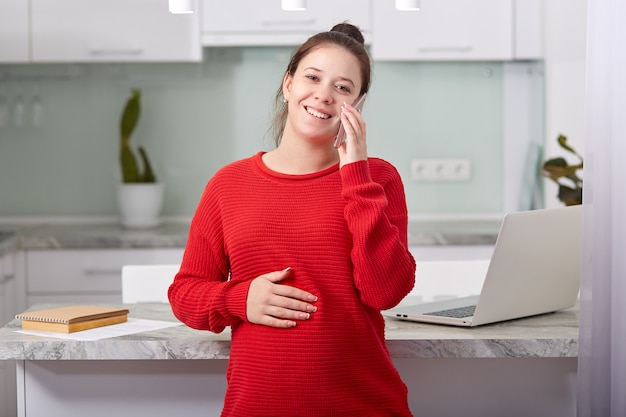 Cheerful future mother has pleasant conversation with close friend, speaks via smart phone, models against kitchen interior, being on maternity leave, spends time at home.