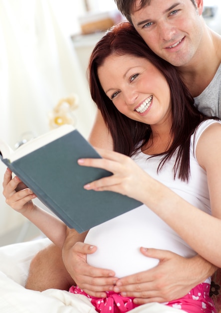 cheerful future parents reading a book sitting on their bed during the morning