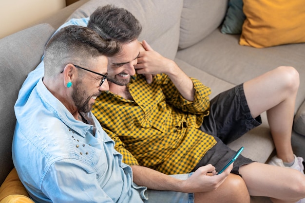 사진 쾌활한 게이 커플은 소파에 앉아 소셜 네트워크를 스크롤하는 영화를 보고 휴대전화를 보고 있다
