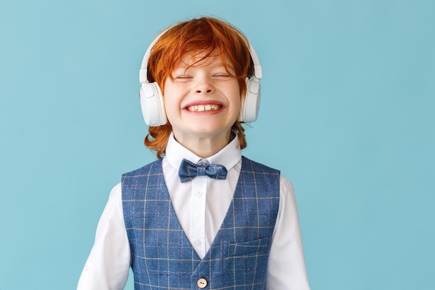Cheerful ginger boy in school uniform with closed eyes smiling\
while listening to music in headphones against blue background