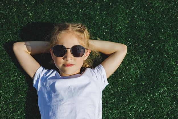 사진 선글라스를 끼고 8살 된 쾌활한 소녀가 공원의 잔디에 누워 있습니다.