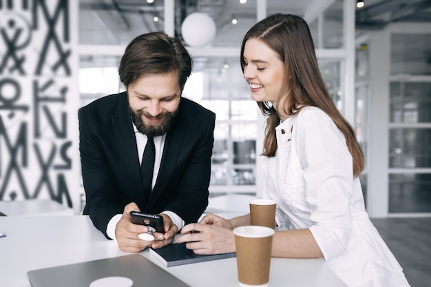 Веселая девушка и бородатый мужчина команда стартапов со смартфоном пьют кофе в офисе