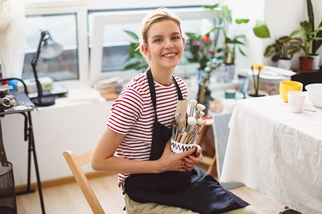 Cheerful girl in black apron and striped t-shirt holding bowl\
with pottery tools in hands happily looking in camera at modern\
pottery studio