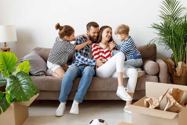 cheerful girl and boy tickling laughing parents while resting on settee near cardboard boxes and plants in new apartment