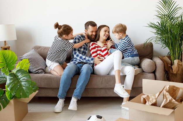 Cheerful girl and boy tickling laughing parents while resting on settee near cardboard boxes and plants in new apartment