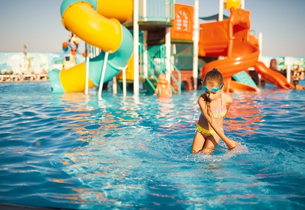 밝은 수영복과 파란색 수영 고글을 쓴 쾌활한 소녀가 물에 손을 넣고 맑은 물이 있는 수영장에서 회전하고 있습니다