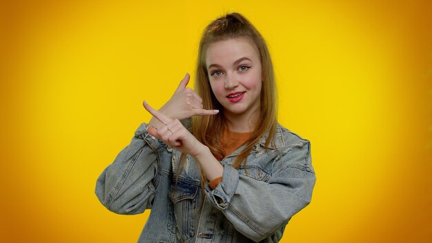 Cheerful girl in denim jacket looking at camera doing phone gesture like says hey you call me back person