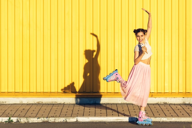 A cheerful girl eats ice cream and rolls on rollers in summer against the background of a yellow wall.