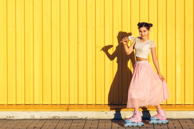 A cheerful girl eats ice cream and rolls on rollers in summer against the of a yellow wall