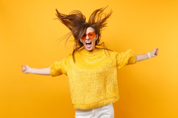 Cheerful girl in fur sweater and heart orange glasses screaming fooling around in studio jumping with flowing hair isolated on yellow background. people sincere emotions, lifestyle. advertising area.