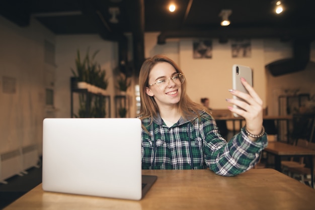 Cheerful girl in glasses and a casual dress sitting in a cafe with a laptop, makes selfie on a smartphone, smiling and posing