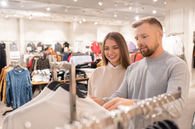 Cheerful girl and her boyfriend looking through jumpers or sweatshirts in casualwear department in the mall or trade center