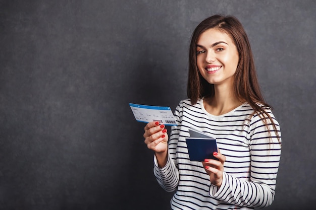 Cheerful girl holding passport plane ticket before grey background indoor travel concept