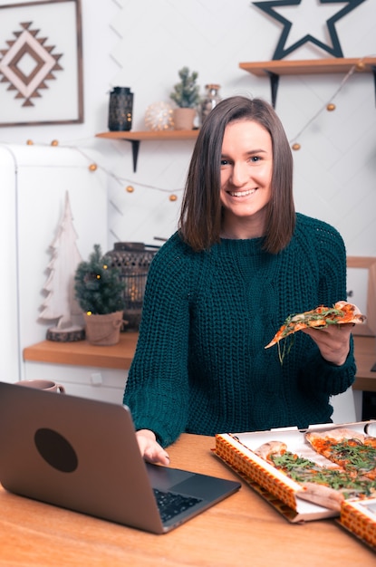 Cheerful girl is sitting in the kitchen, at her laptop near an ordered pizza.