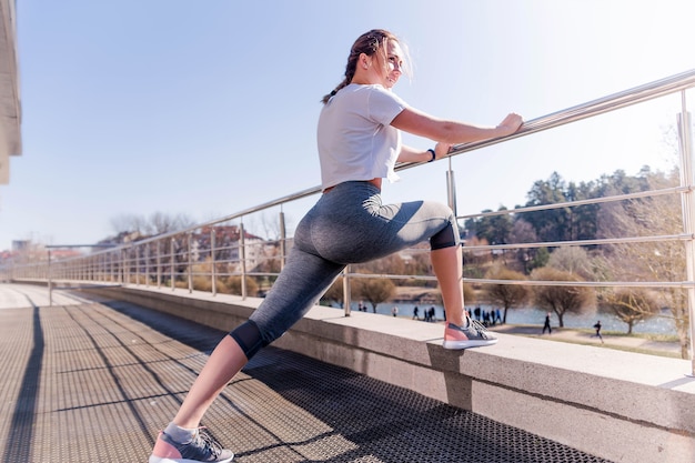 A cheerful girl is suitable for a morning run. Young woman in a white T-shirt and gray leggings, leaning on a railing and stretching legs and ankles. Warm up concept