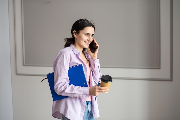 A cheerful girl is talking on the phone and holding a folder with documents and a cup