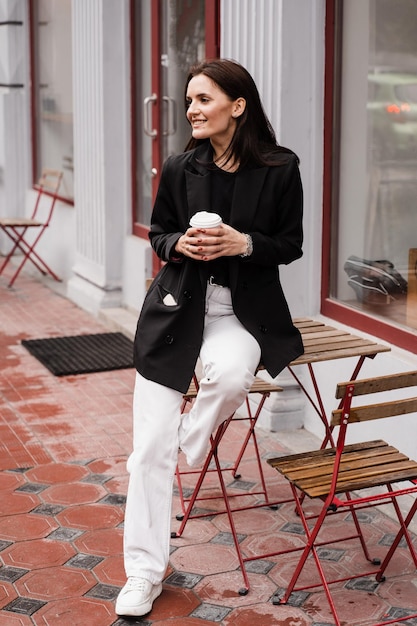 Cheerful girl is weared casual business style is posing near cafe Confident young business woman with cup of coffee is posing outside of cafe on terrace