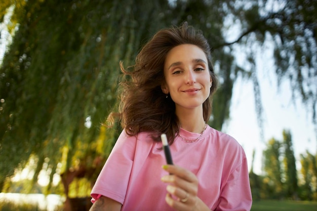 Cheerful girl in a pink t-shirt smile, holding an electronic cigarette, in the park.