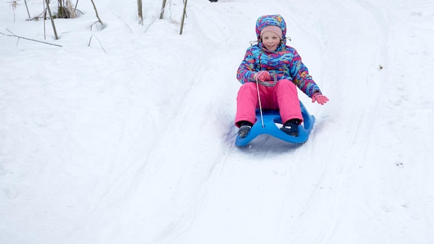 白い日当たりの良い冬の山の風景の中、雪に覆われたそりの道を下り坂でそりに乗っている陽気な女の子