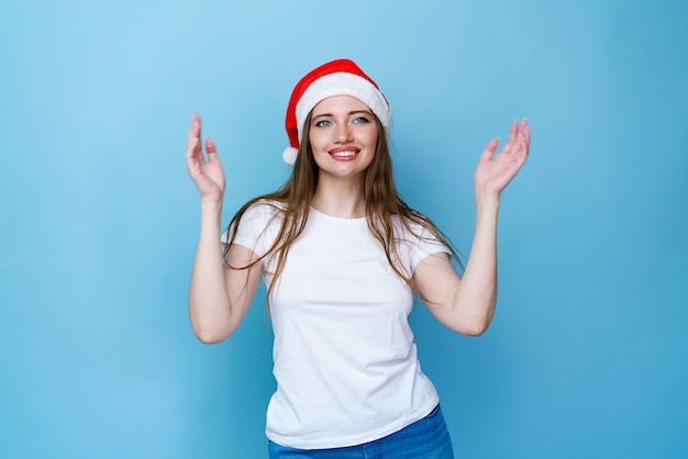 Cheerful girl in santa hat posing and smiling with toothy smile on blue background in a white tshirt...