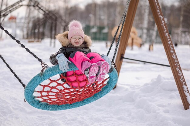 Cheerful girl swings on wicker swing for walk outdoors on frosty winter day in park