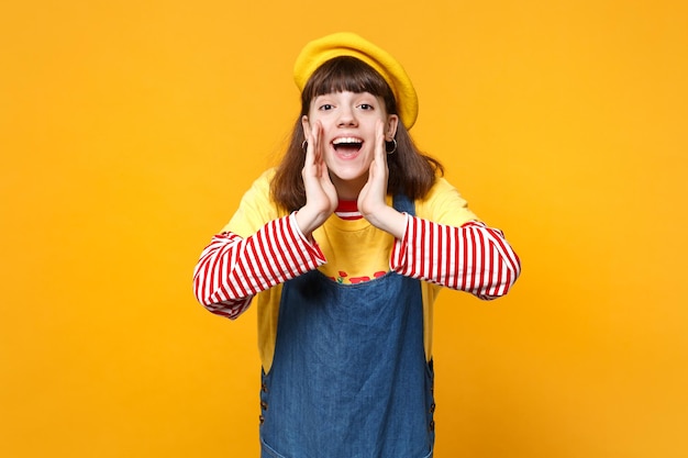 Cheerful girl teenager in french beret, denim sundress whispers gossip and tells secret with hand gesture isolated on yellow background. People sincere emotions, lifestyle concept. Mock up copy space.