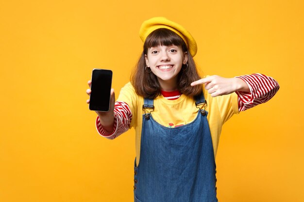 Cheerful girl teenager in french beret pointing index finger on mobile phone with blank empty screen isolated on yellow wall background. People sincere emotions, lifestyle concept. Mock up copy space.