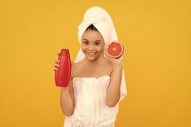 Cheerful girl in towel with grapefruit shampoo bottle on yellow background