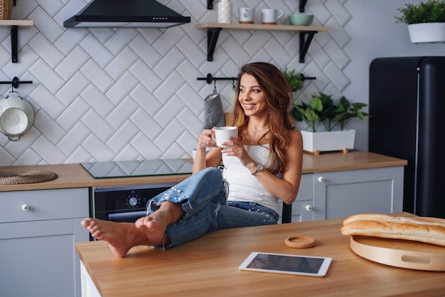 Cheerful girl in white t-shirt using tablet computer while relaxing in a chair at home in cozy kitchen.