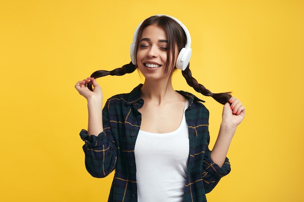 Cheerful girl with headphones enjoy sound of music holds her pony tails with closed eyes above yellow wall