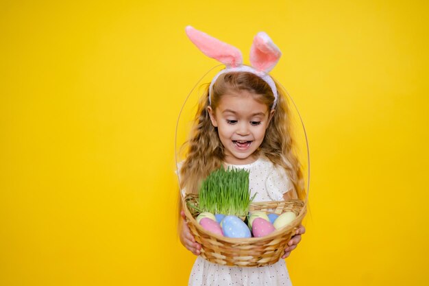 A cheerful girl with rabbit ears on her head with a basket of colored eggs in her hands on a yelow background Funny crazy happy baby Easter to the child High quality photo