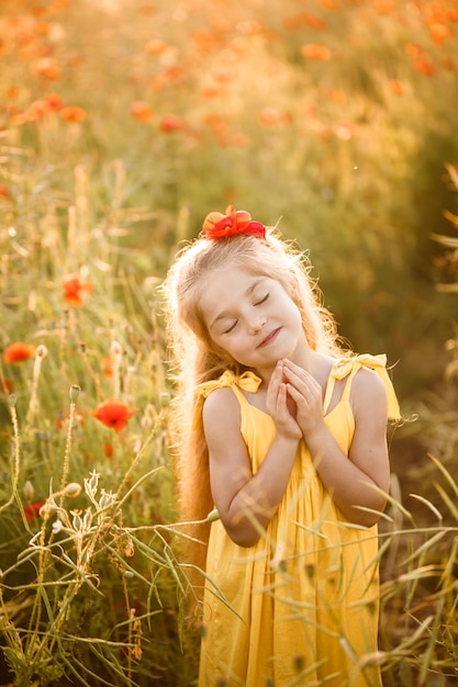 Cheerful girl with a smile on her face in blooming field poppies. Red flowers. A blonde in a yellow short dress and a hat.