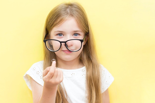 A cheerful girl on a yellow background wearing glasses and holding a contact lens. The concept of vision problems in school children.