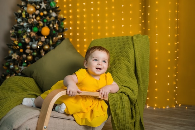 A cheerful girl in a yellow dress is sitting in a rocking chair with Christmas tree