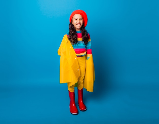 A cheerful girl in a yellow raincoat, red beret and rubber boots jumps with outstretched arms and looks at the camera.