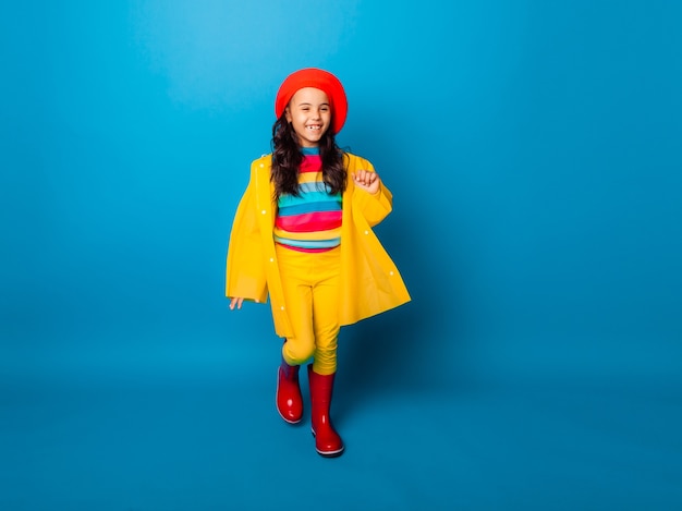 A cheerful girl in a yellow raincoat, red beret and rubber boots jumps with outstretched arms and looks at the camera.