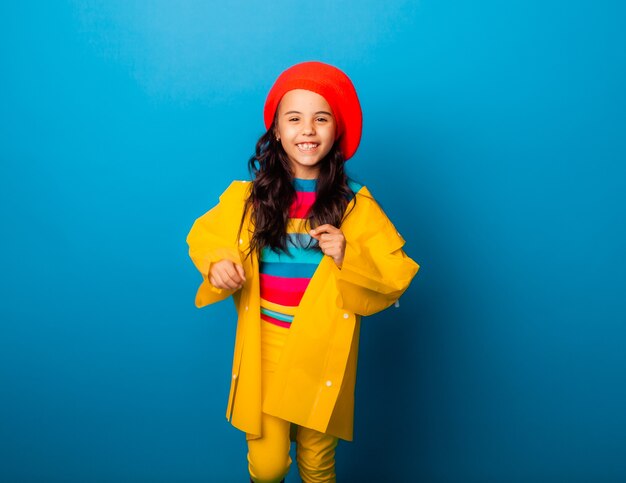 A cheerful girl in a yellow raincoat, red beret and rubber boots jumps with outstretched arms and looks at the camera.