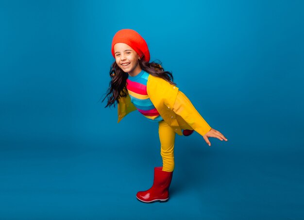 A cheerful girl in a yellow raincoat, red beret and rubber boots jumps with outstretched arms and looks at the camera.