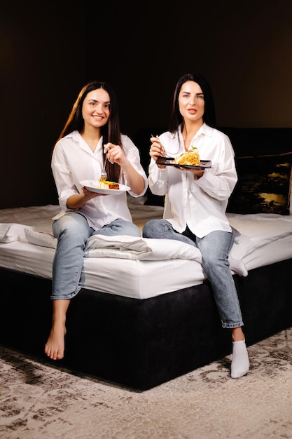 Cheerful girls eating cakes sitting on bedroom in apartment