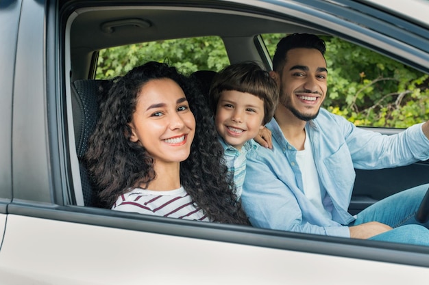 Cheerful glad millennial arab mother and father with kid go on vacation by auto and look at camera at open window