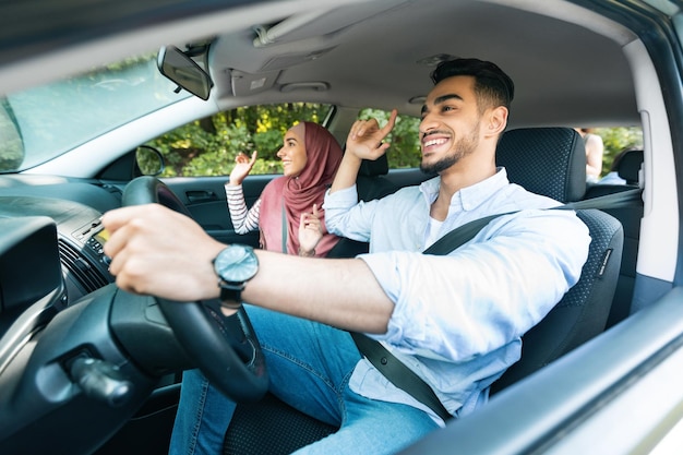 Cheerful glad young arab man driving with female in hijab ride in car couple enjoy music and dance