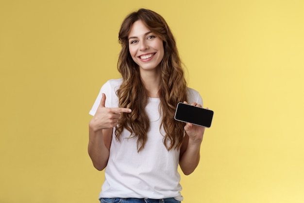 Веселая симпатичная дружелюбная беззаботная молодая девушка держит смартфон горизонтально указывая указательным пальцем на экране мобильного телефона, широко улыбаясь, и представляет потрясающую игровую подставку для приложений на желтом фоне.