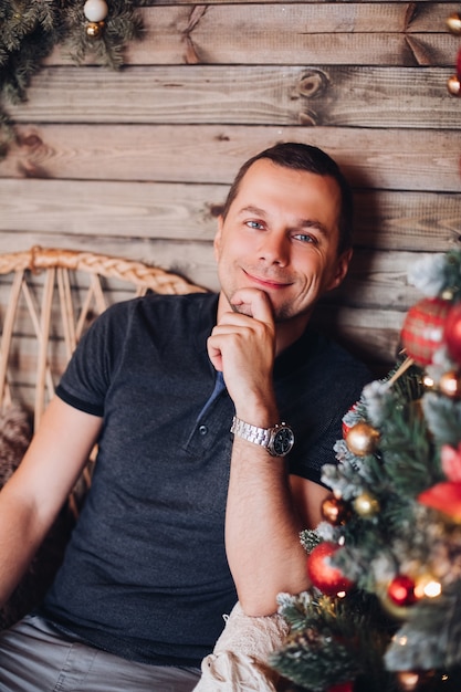 Веселый красивый мужчина в футболке, улыбаясь в камеру, отдыхая у деревянной стены и рождественского дерева.
