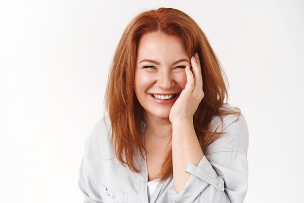 Cheerful good-looking redhead mature woman carefree not care aging taking care skin no wrinkles touch cheek pleased laughing joyfully enjoy squinting happily standing white wall