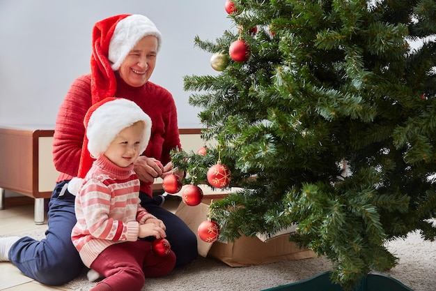 陽気な祖母と彼女のかわいい孫娘の女の子がクリスマスツリーを飾ります。祖母と小さな子供が屋内の木の近くで楽しんでいます。自宅の部屋にいる愛する家族。