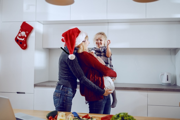 陽気な祖母、妊娠中の女性、キッチンで抱き締める少女。キッチンカウンターには野菜とラップトップがあります。クリスマスコンセプトの充実した時間。