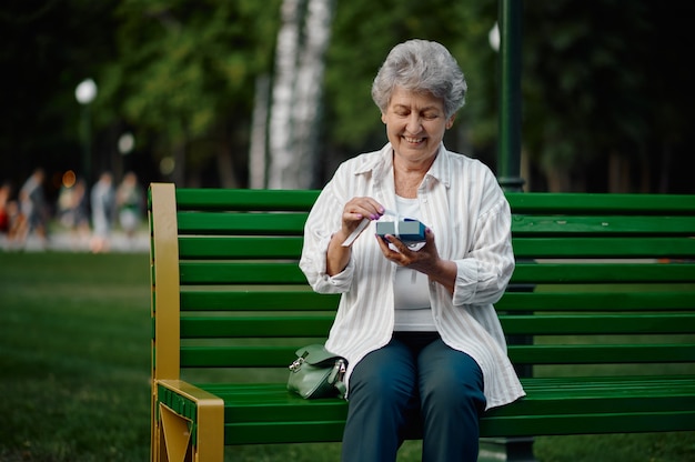 Cheerful granny opens a gift box on the bench in summer park. Aged people lifestyle. Pretty grandmother having fun outdoors, old female person on nature