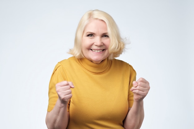 Cheerful granny with toothy smile raised hands and showing successful sign