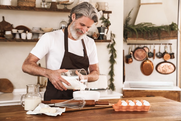 cheerful gray-haired man in apron cooking dough for pastry at cozy kitchen